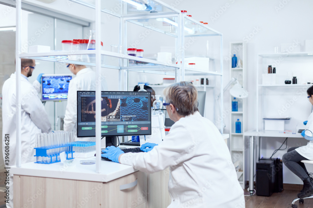 Doctor in chemistry and her multiethnic teamn engineering drug. Senior scientist in pharmaceuticals laboratory doing genetic research wearing lab coat with team in the background. Doctor in chemistry and her multiethnic teamn engineering drug. Senior scientist in pharmaceuticals laboratory doing genetic research wearing lab coat with team in the background.
