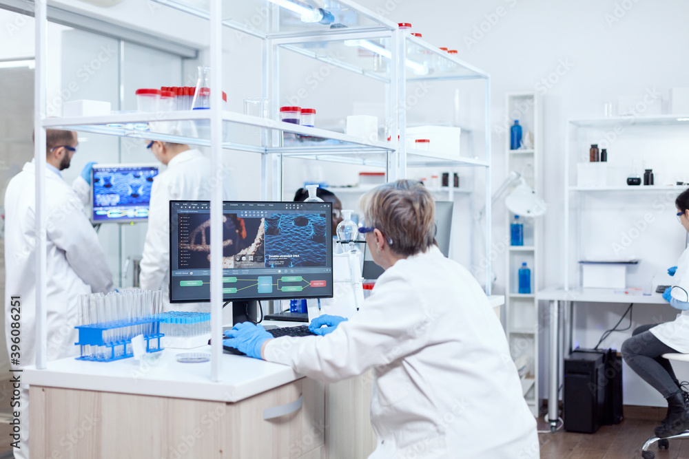 Doctor in chemistry and her multiethnic teamn engineering drug. Senior scientist in pharmaceuticals laboratory doing genetic research wearing lab coat with team in the background.