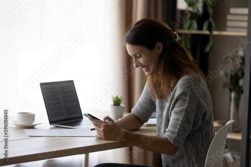 Side view smiling millennial woman excited by reading email message with good news on mobile telephone, distracted from online work or distant study on computer, sitting at home office workplace.