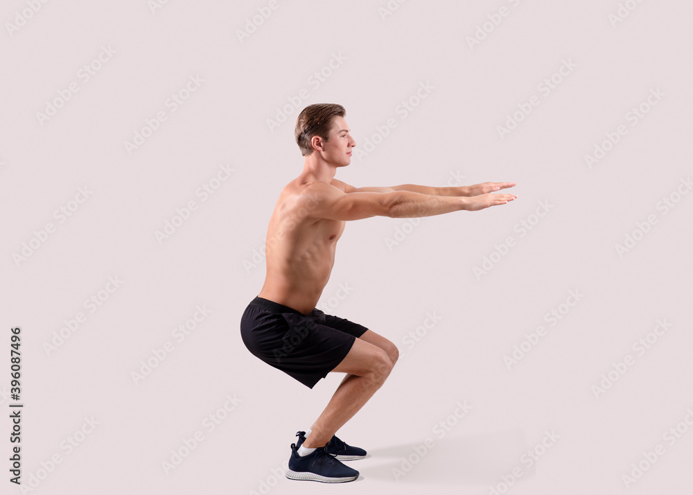 Side view of strong young guy doing squats on light studio background, full length portrait