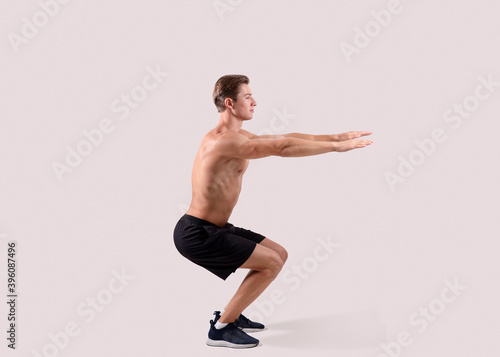 Side view of strong young guy doing squats on light studio background, full length portrait