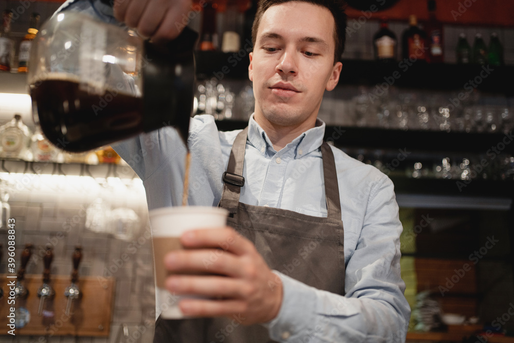 a staff member in a small restaurant works in an apron and shirt. Barista waiter prepares sells coffee Americano cappuccino with a takeaway in the cafe. pours a paper Cup from the teapot into a Cup.
