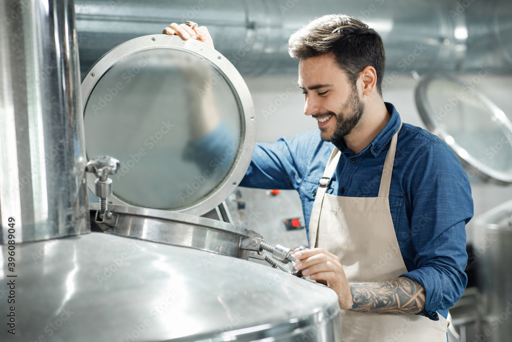Smiling male worker or owner in apron opens lid of large boiler for brewing beer and checking fermentation Smiling male worker or owner in apron opens lid of large boiler for brewing beer and checking fermentation