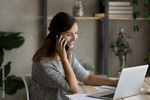 Smiling young businesswoman holding mobile call conversation with clients or colleagues, working on computer at home office, discussing project or brainstorming ideas, distant workday concept.