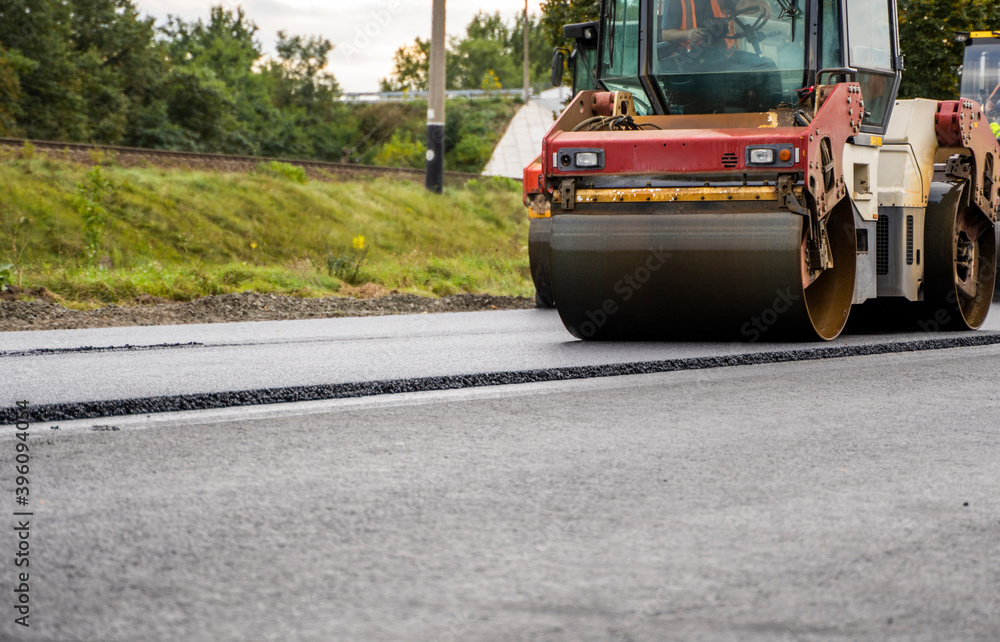 Fototapeta premium Asphalt road roller with heavy vibration roller compactor press new hot asphalt on the roadway on a road construction site. Heavy Vibration roller at asphalt pavement working. Repairing.
