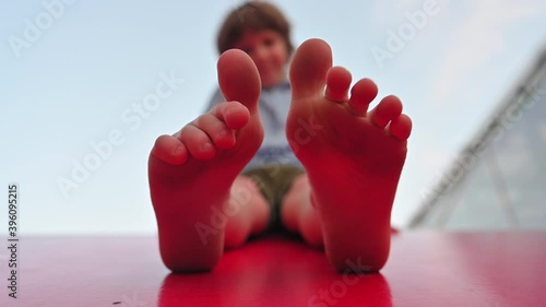 Boy plays with his fingers on his feet sitting on the red roof of a children's house close