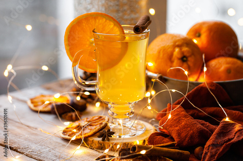 Delicious hot winter drink in glass mug: spicy tea with cinnamon, anise, orange. Citrus is a natural antiviral remedy. Cozy home holiday atmospehere with christmas lights on background.  Close up photo