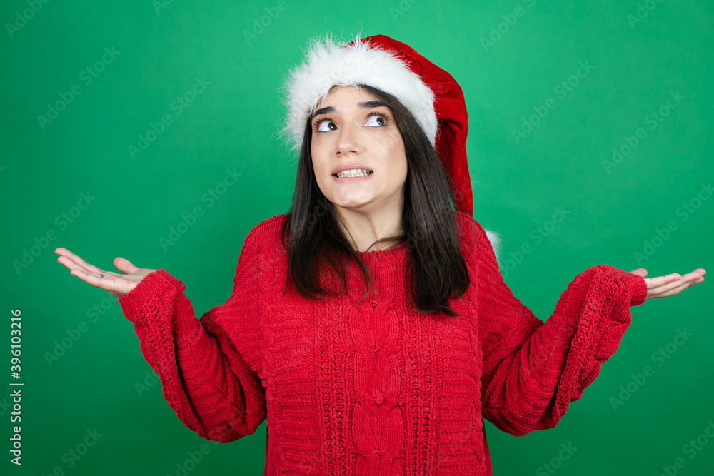 Young beautiful woman wearing Christmas Santa hat over isolated green background clueless and confused expression with arms and hands raised Young beautiful woman wearing Christmas Santa hat over isolated green background clueless and confused expression with arms and hands raised