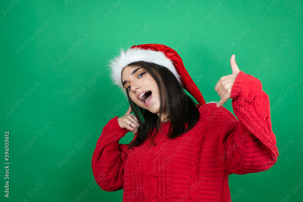 Young beautiful woman wearing Christmas Santa hat over isolated green background doing the “call me” gesture with her hands. Young beautiful woman wearing Christmas Santa hat over isolated green background doing the “call me” gesture with her hands.