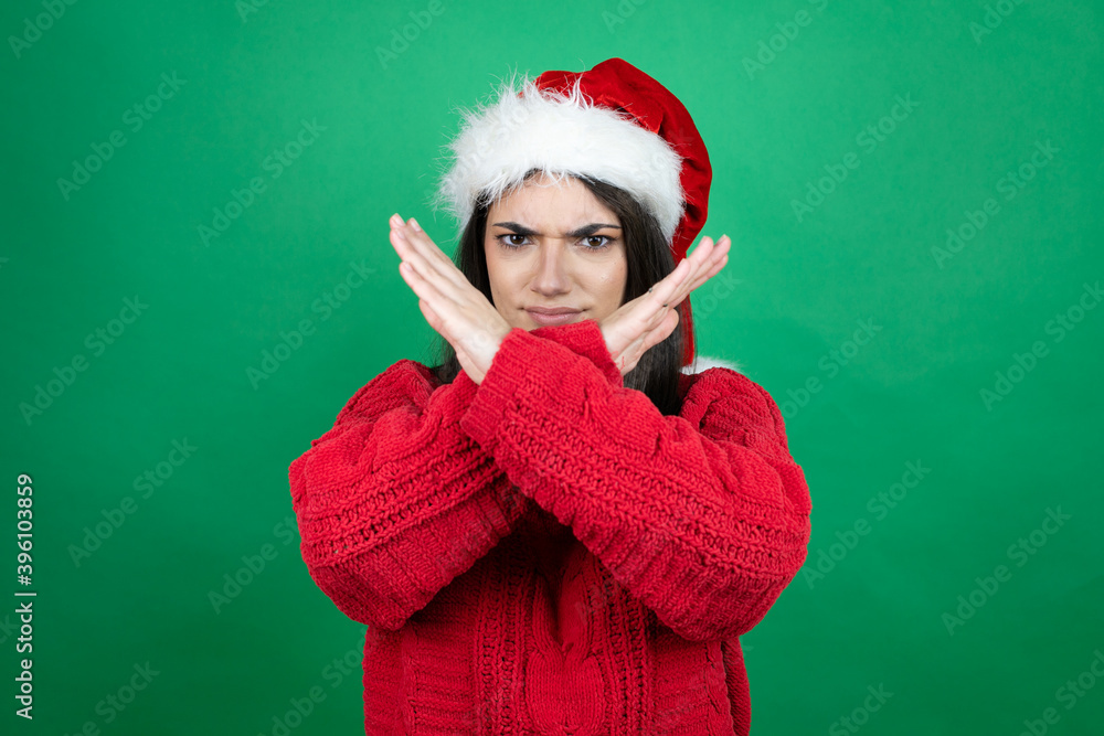 Young beautiful woman wearing Christmas Santa hat over isolated green background Rejection expression crossing arms doing negative sign, angry face Young beautiful woman wearing Christmas Santa hat over isolated green background Rejection expression crossing arms doing negative sign, angry face