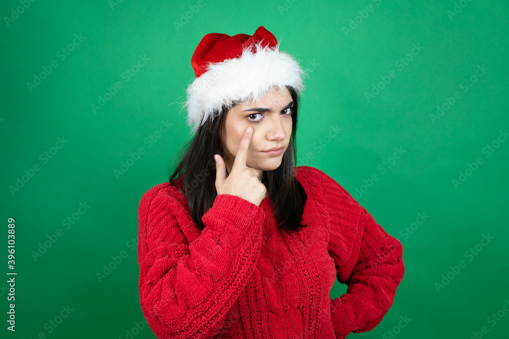Young beautiful woman wearing Christmas Santa hat over isolated green background Pointing to the eye watching you gesture, suspicious expression Young beautiful woman wearing Christmas Santa hat over isolated green background Pointing to the eye watching you gesture, suspicious expression