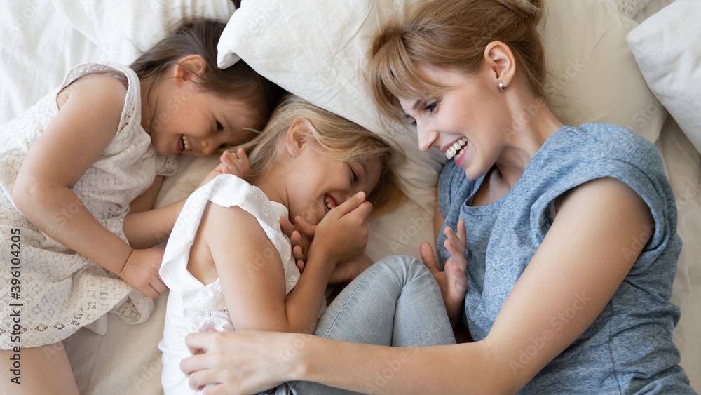 Top view overjoyed mother and two little daughters lying in cozy bed together, hugging and cuddling, laughing mum and adorable kids having fun, tickling, enjoying leisure time at home Top view overjoyed mother and two little daughters lying in cozy bed together, hugging and cuddling, laughing mum and adorable kids having fun, tickling, enjoying leisure time at home