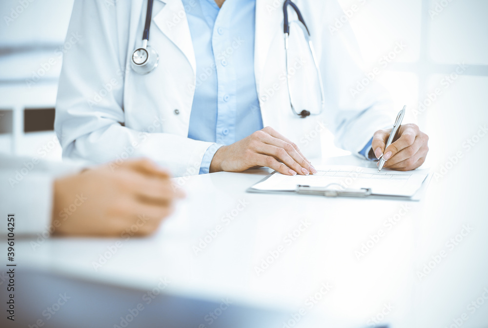 Unknown woman-doctor and female patient sitting and talking at medical examination in clinic, close-up. Therapist wearing blue blouse is filling up medication history record. Medicine concept Unknown woman-doctor and female patient sitting and talking at medical examination in clinic, close-up. Therapist wearing blue blouse is filling up medication history record. Medicine concept