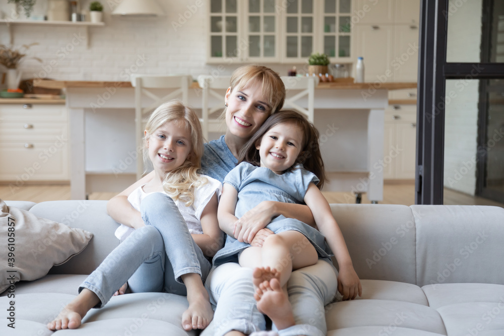 Family portrait smiling young woman hugging two adorable daughters, happy beautiful mother with kids looking at camera, sitting on cozy couch in modern living room, posing for photo, having fun Family portrait smiling young woman hugging two adorable daughters, happy beautiful mother with kids looking at camera, sitting on cozy couch in modern living room, posing for photo, having fun