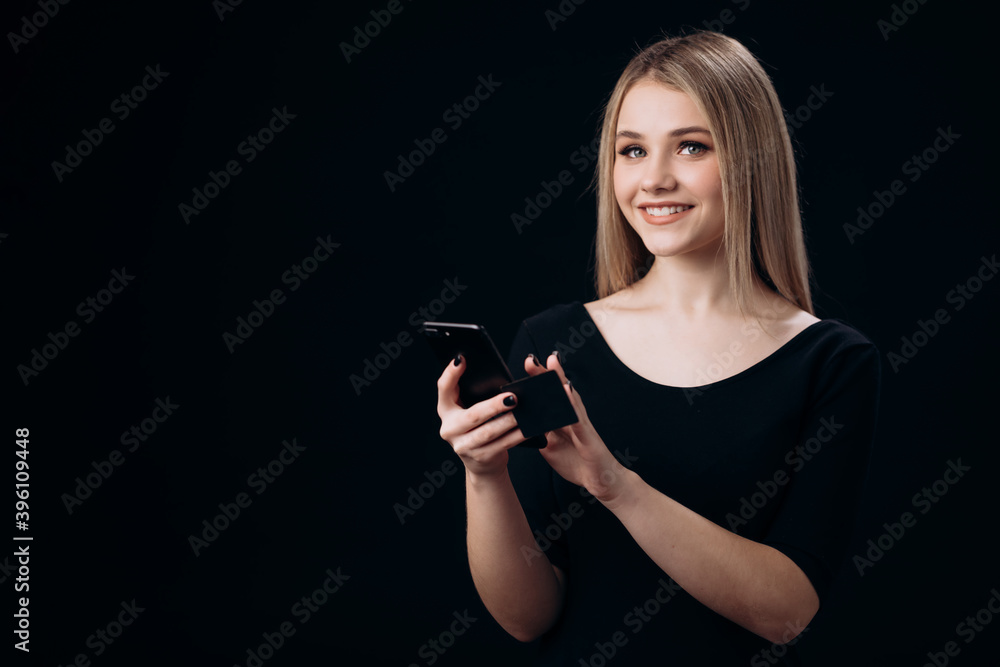 Portrait of smiling young woman with blond hair using smartphone and credit card for online shopping. Isolated over black studio background. Portrait of smiling young woman with blond hair using smartphone and credit card for online shopping. Isolated over black studio background.