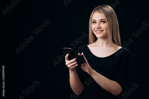 Portrait of smiling young woman with blond hair using smartphone and credit card for online shopping. Isolated over black studio background.