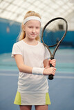 Cute blond little girl in activewear holding tennis racket against left shoulder