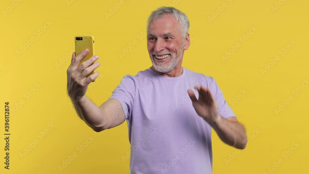 Smiling elderly gray-haired mustache bearded man in t-shirt isolated on yellow background studio. People lifestyle concept. Talking mobile phone making video call greeting with hand showing thumb up