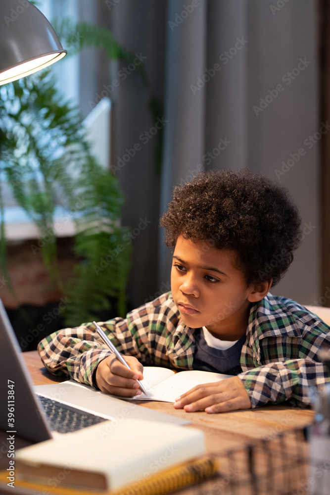Serious African schoolboy making notes in copybook during online lesson Serious African schoolboy making notes in copybook during online lesson