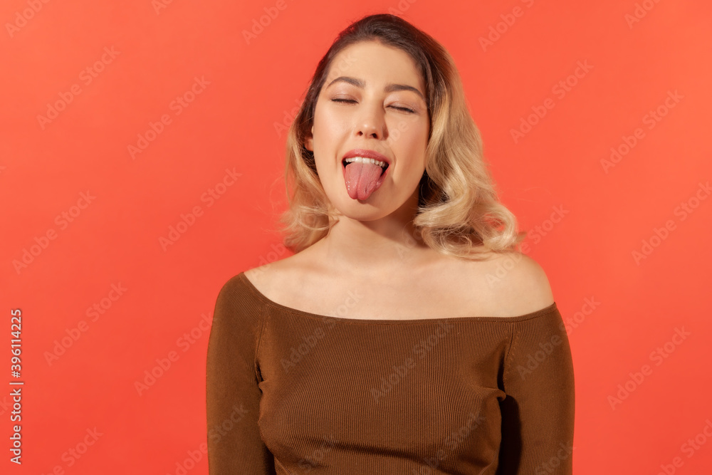 Portrait of funny disobedient young woman with blonde hair keeping eyes closed and demonstrating tongue, behaving naughty unruly, childish mood. Indoor studio shot isolated on red background Portrait of funny disobedient young woman with blonde hair keeping eyes closed and demonstrating tongue, behaving naughty unruly, childish mood. Indoor studio shot isolated on red background