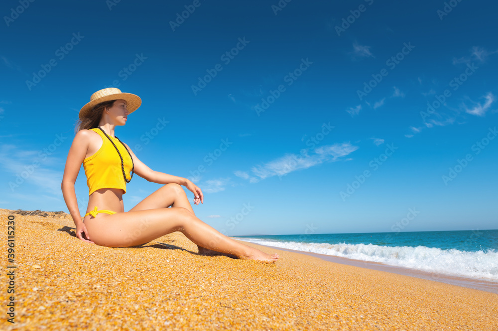 Beautiful slim and attractive caucasian young woman in a yellow swimsuit and top wearing a straw hat enjoys the sea while sitting on the sand on the sea beach on a sunny day.