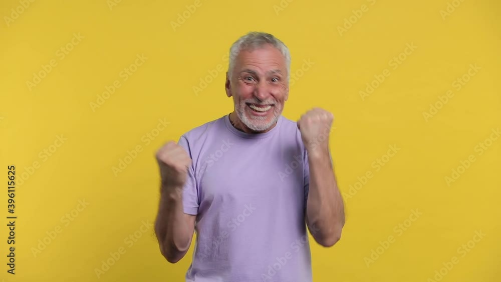Shocked happy joyful elderly gray-haired mustache bearded man in basic violet t-shirt isolated on yellow color background studio. People lifestyle concept. Doing winner gesture clenching fists dancing Shocked happy joyful elderly gray-haired mustache bearded man in basic violet t-shirt isolated on yellow color background studio. People lifestyle concept. Doing winner gesture clenching fists dancing
