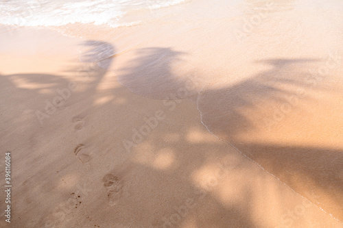 Tropical beach sand and waves with shadows of coconut palm tree leaves and human footprints. Travel and vacations concept background.