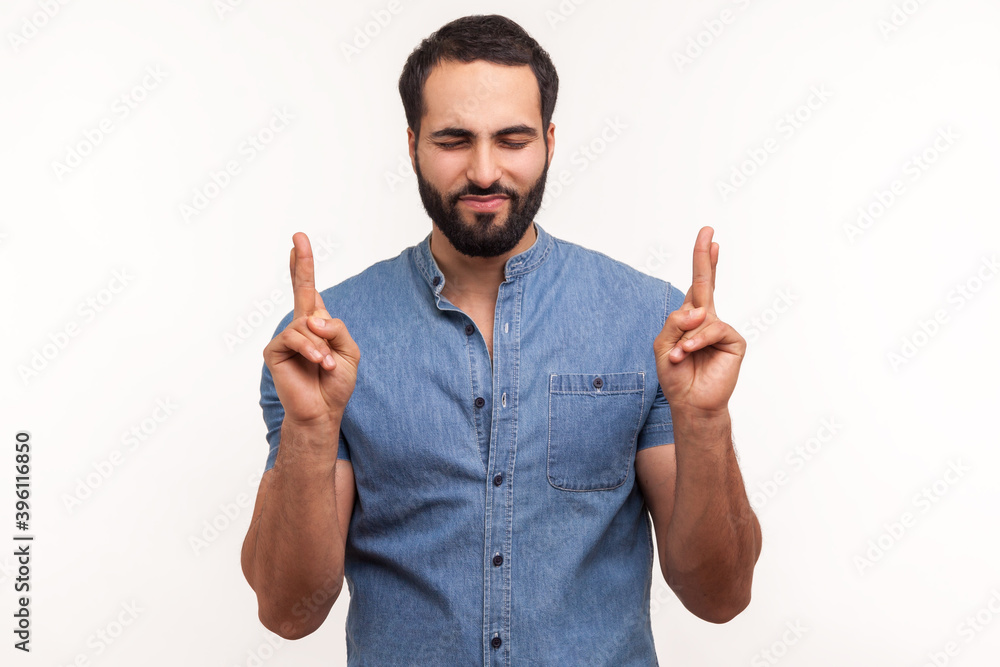 Hopeful dreamy man with beard in blue shirt crossing fingers and closing eyes making wish, believing in miracle, hope in success. Indoor studio shot isolated on white background Hopeful dreamy man with beard in blue shirt crossing fingers and closing eyes making wish, believing in miracle, hope in success. Indoor studio shot isolated on white background