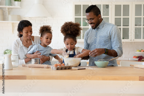Family recipe. Happy african family mother, father and younger son watching with interest affection elder daughter kneading dough, black parents and small children baking holiday cake at home together