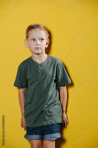 Portrait of a uneasy ten year old boy standing in front of a yellow wall, leaning on it.