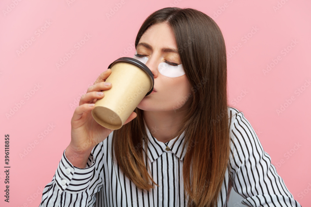 Tired sleepy woman with brown hair in striped shirt doing beauty procedure wearing eye patches and drinking coffee from paper cup, lack of energy. Indoor studio shot isolated on pink background Tired sleepy woman with brown hair in striped shirt doing beauty procedure wearing eye patches and drinking coffee from paper cup, lack of energy. Indoor studio shot isolated on pink background