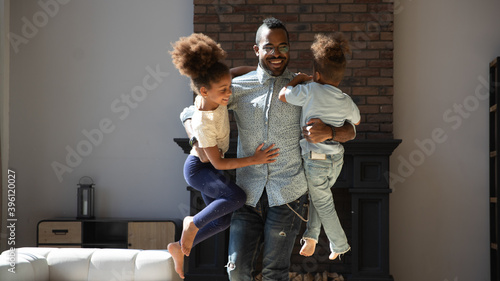 Daddy is the strongest. Happy laughing african father or male babysitter spending time at home with two active kids elder preteen sister and little brother cuddling holding in air playing funny games