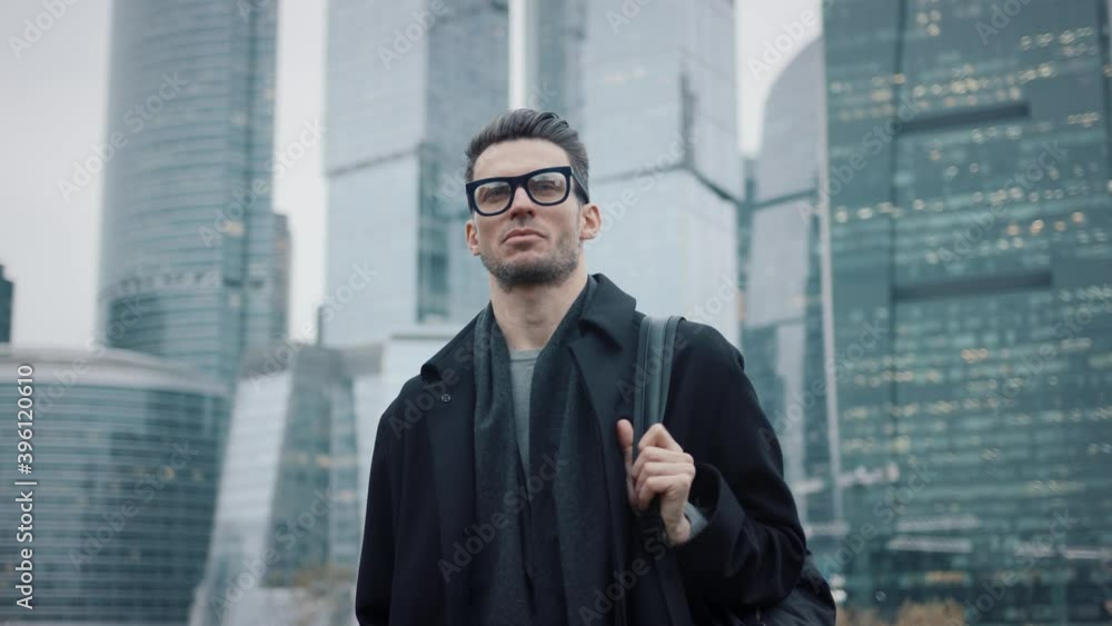 Man in black coat slow motion walking on background of business city skyscrapers in the evening, lights in the windows. Gimbal shot of young businessman in eyeglasses Man in black coat slow motion walking on background of business city skyscrapers in the evening, lights in the windows. Gimbal shot of young businessman in eyeglasses