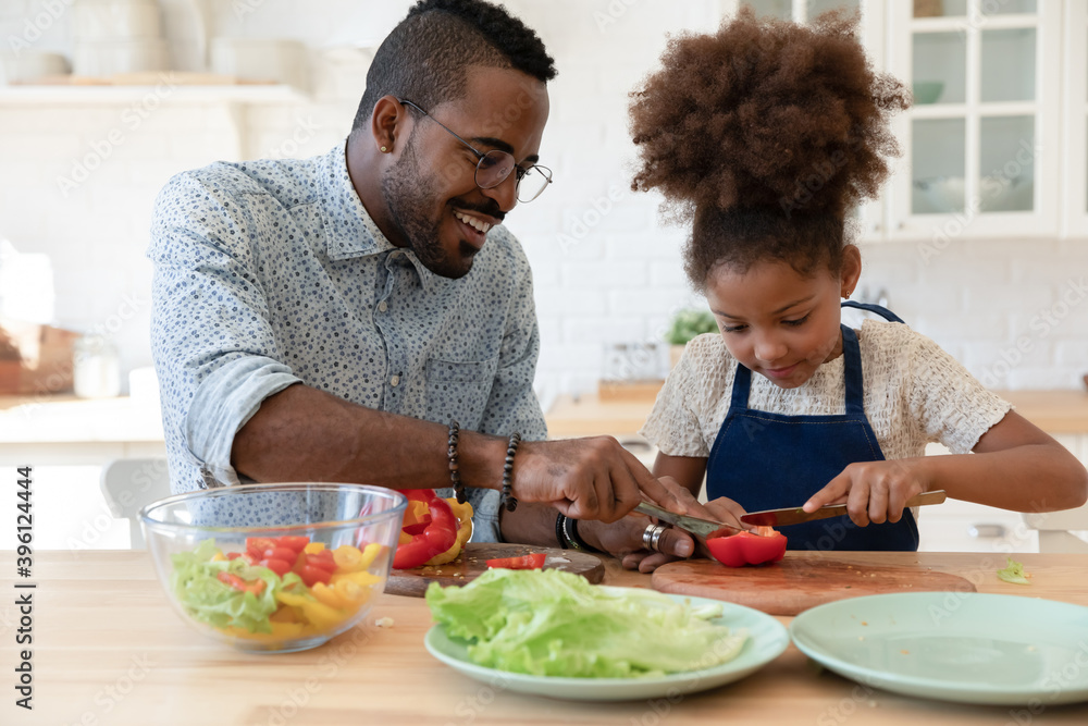 Assisting daddy. Happy smiling single african black dad and small tween daughter engaged in easy cooking together spending time at kitchen talking preparing healthy vegan lunch of lettuce and pepper Assisting daddy. Happy smiling single african black dad and small tween daughter engaged in easy cooking together spending time at kitchen talking preparing healthy vegan lunch of lettuce and pepper