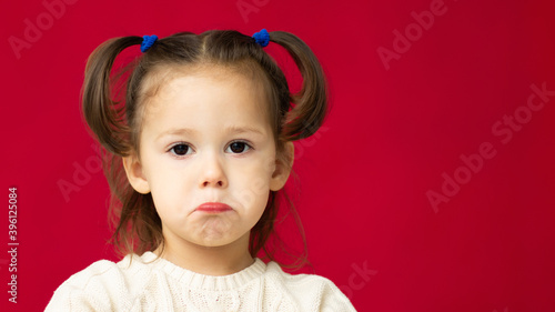 The face of a sad little cute girl 2-4 in a white knitted dress on a red background.