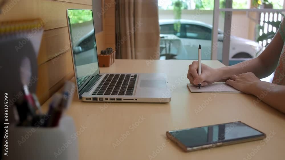 asian woman in casual wear, typing laptop computer and write down the memo notes on post-it paper. female wooden desk working from home gathering information from internet. asian woman in casual wear, typing laptop computer and write down the memo notes on post-it paper. female wooden desk working from home gathering information from internet.