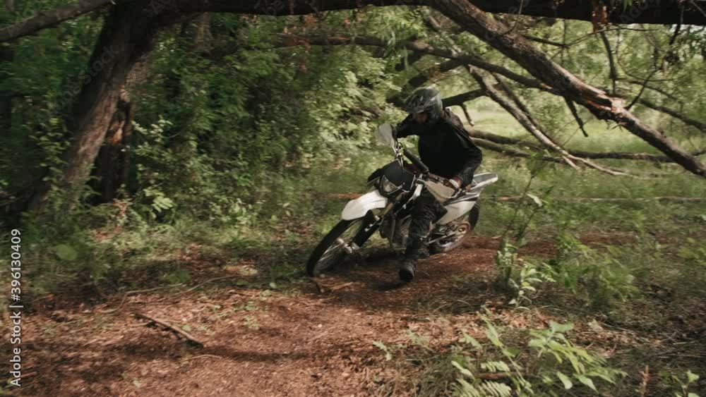 Tracking shot of men in helmets and gear racing on motorcycles along dirt road and turning into forest with lush greenery Tracking shot of men in helmets and gear racing on motorcycles along dirt road and turning into forest with lush greenery