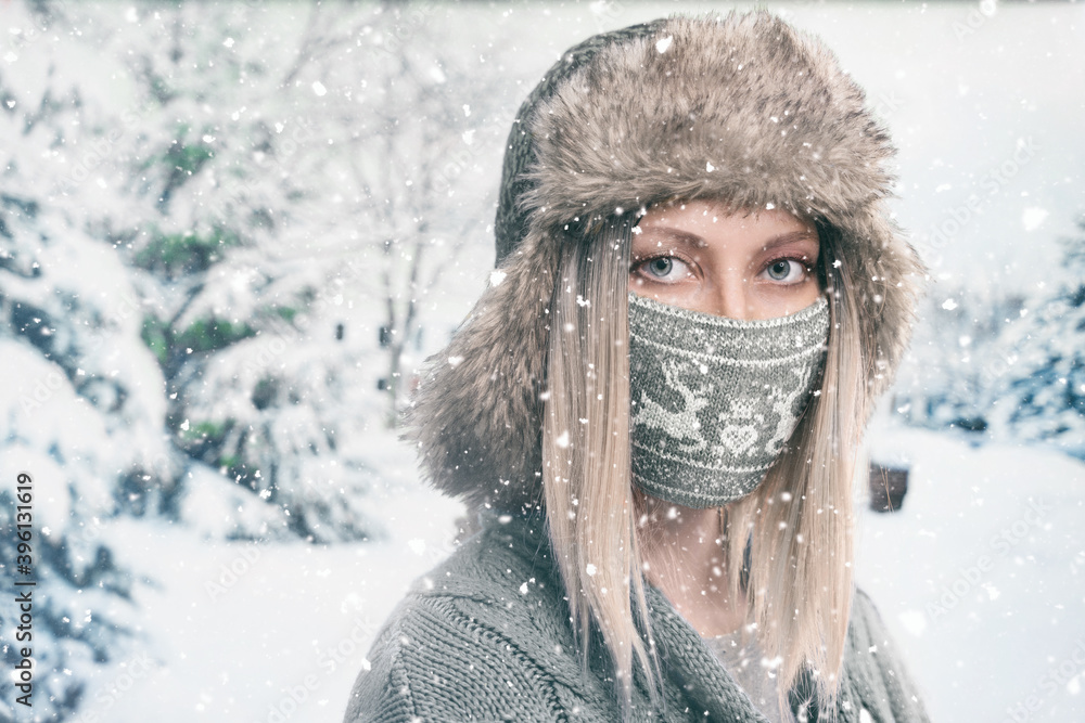 Portrait of a blonde girl in a hat with earflaps and a knitted face mask with deers on a winter background with falling snow Portrait of a blonde girl in a hat with earflaps and a knitted face mask with deers on a winter background with falling snow