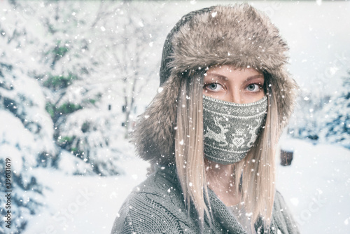 Portrait of a blonde girl in a hat with earflaps and a knitted face mask with deers on a winter background with falling snow