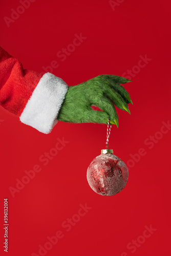 A green hairy hand in a Santa suit holds a red Christmas ball on a red background photo