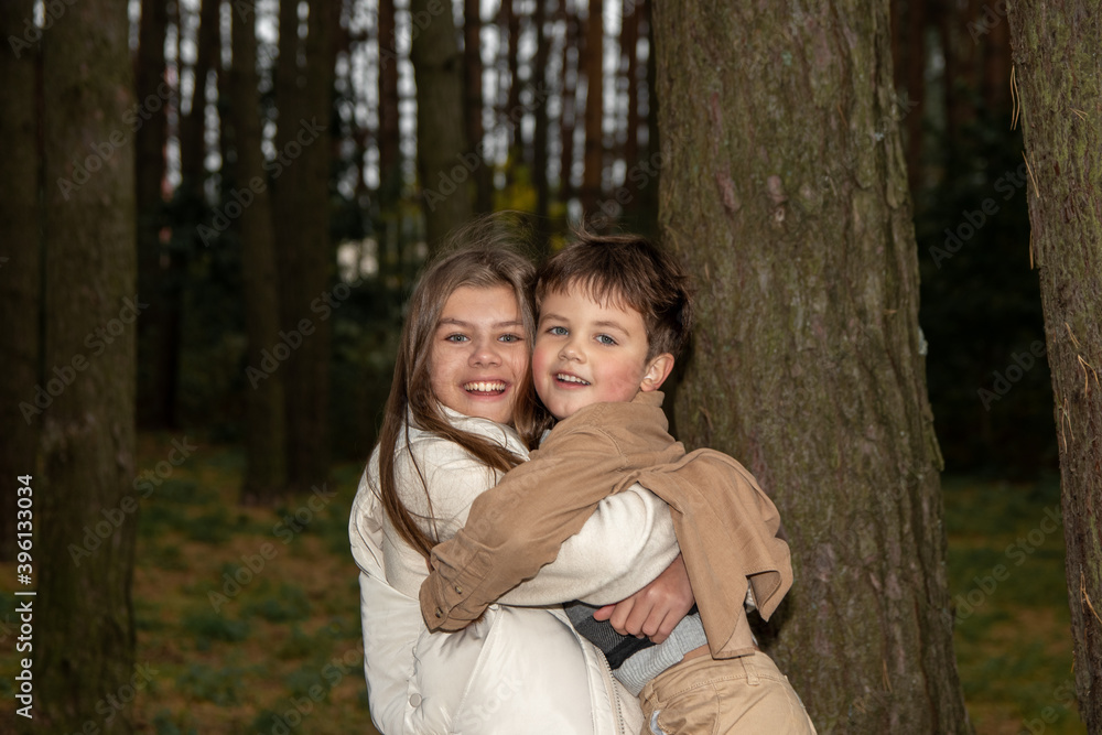 A teenage girl in a light dress hugs her little brother. Children are happy and laugh among the trees in forest. Real childhood and family happiness in the middle of nature. Close-up photo