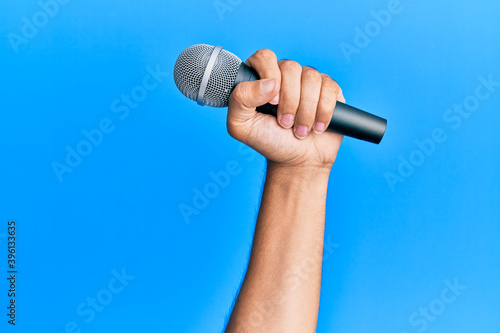 Hand of hispanic man holding microphone over isolated blue background.