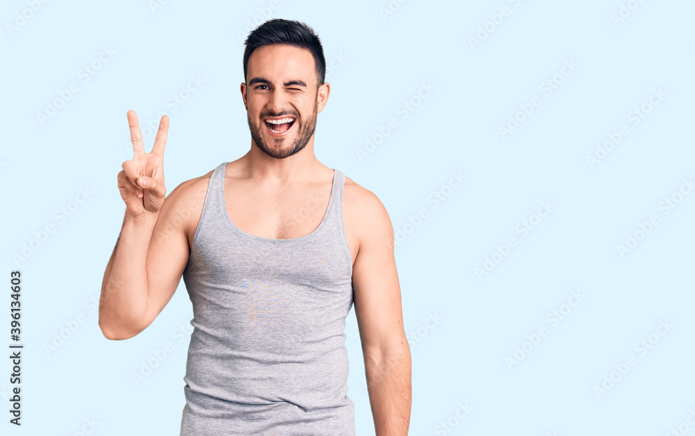 Young handsome man wearing swimwear and sleeveless t-shirt smiling with happy face winking at the camera doing victory sign with fingers. number two. Young handsome man wearing swimwear and sleeveless t-shirt smiling with happy face winking at the camera doing victory sign with fingers. number two.