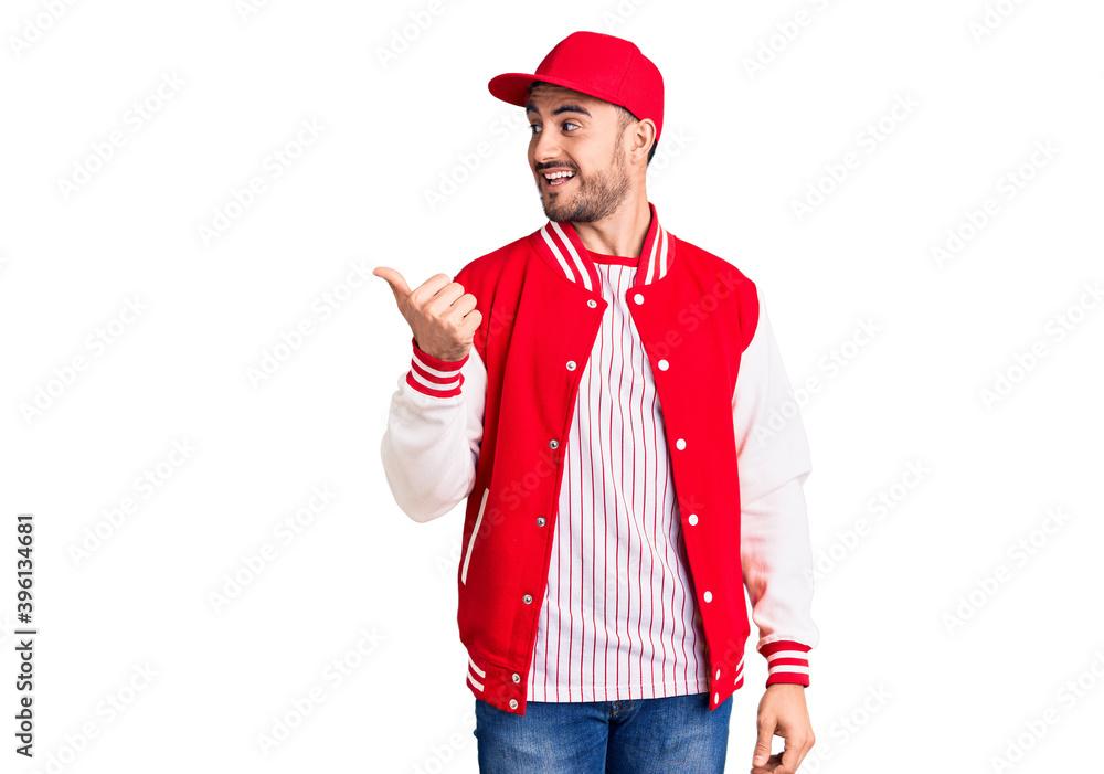 Young handsome man wearing baseball jacket and cap smiling with happy face looking and pointing to the side with thumb up. Young handsome man wearing baseball jacket and cap smiling with happy face looking and pointing to the side with thumb up.