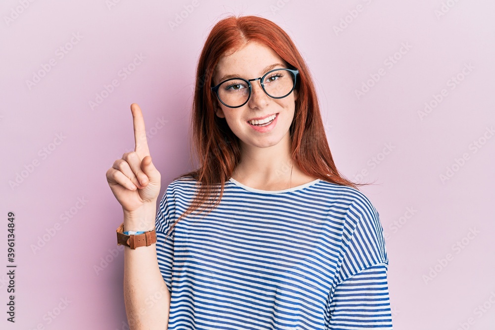 Young red head girl wearing casual clothes and glasses smiling with an idea or question pointing finger up with happy face, number one Young red head girl wearing casual clothes and glasses smiling with an idea or question pointing finger up with happy face, number one