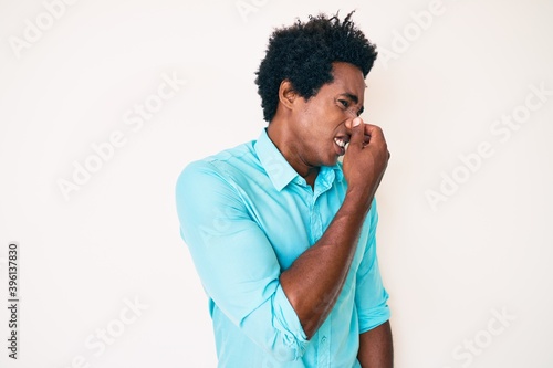 Handsome african american man with afro hair wearing casual clothes smelling something stinky and disgusting, intolerable smell, holding breath with fingers on nose. bad smell