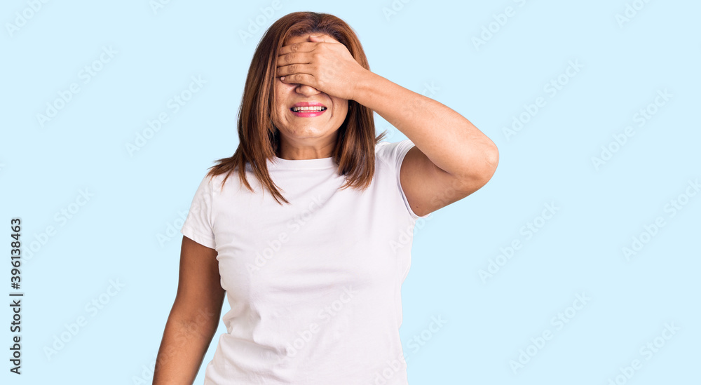 Middle age latin woman wearing casual white tshirt smiling and laughing with hand on face covering eyes for surprise. blind concept. Middle age latin woman wearing casual white tshirt smiling and laughing with hand on face covering eyes for surprise. blind concept.