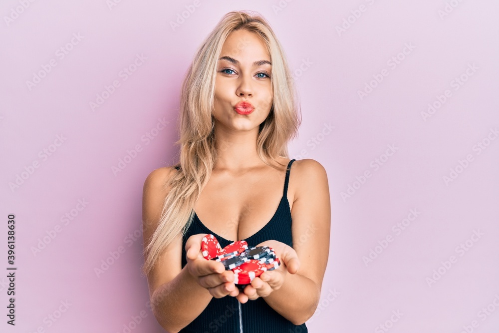 Beautiful caucasian blonde girl holding casino chips looking at the camera blowing a kiss being lovely and sexy. love expression. Beautiful caucasian blonde girl holding casino chips looking at the camera blowing a kiss being lovely and sexy. love expression.