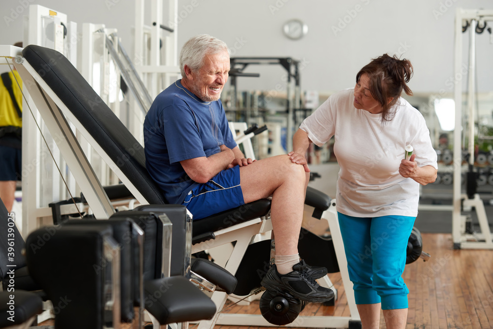 Obraz premium Unhappy senior man with severe pain in knee. Stressed elderly man injured leg at fitness club. Sport trauma concept.