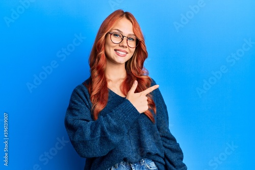 Young beautiful redhead woman wearing casual sweater and glasses over blue background cheerful with a smile of face pointing with hand and finger up to the side with happy and natural expression